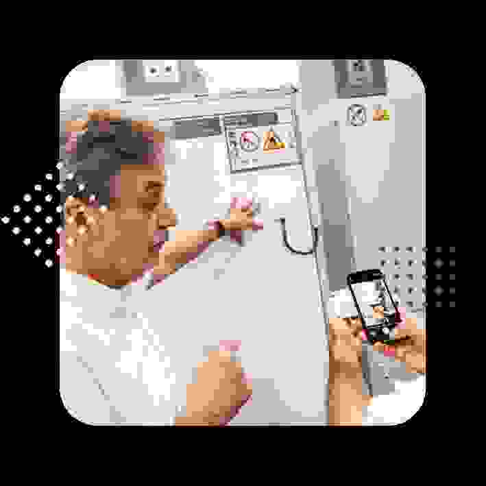 Ein Mann zeigt auf ein Gerät, während eine andere Person mit einem Smartphone ein Foto macht. Beide stehen in einem technischen Umfeld.
