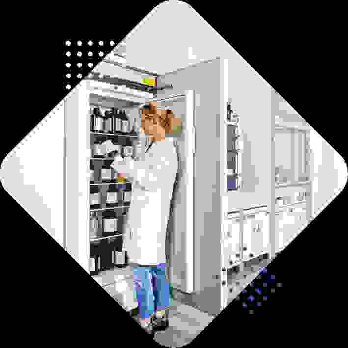 Eine Laborantin in einem weißen Laborkittel steht vor einem Kühlschrank, der mit verschiedenen Chemikalien gefüllt ist.