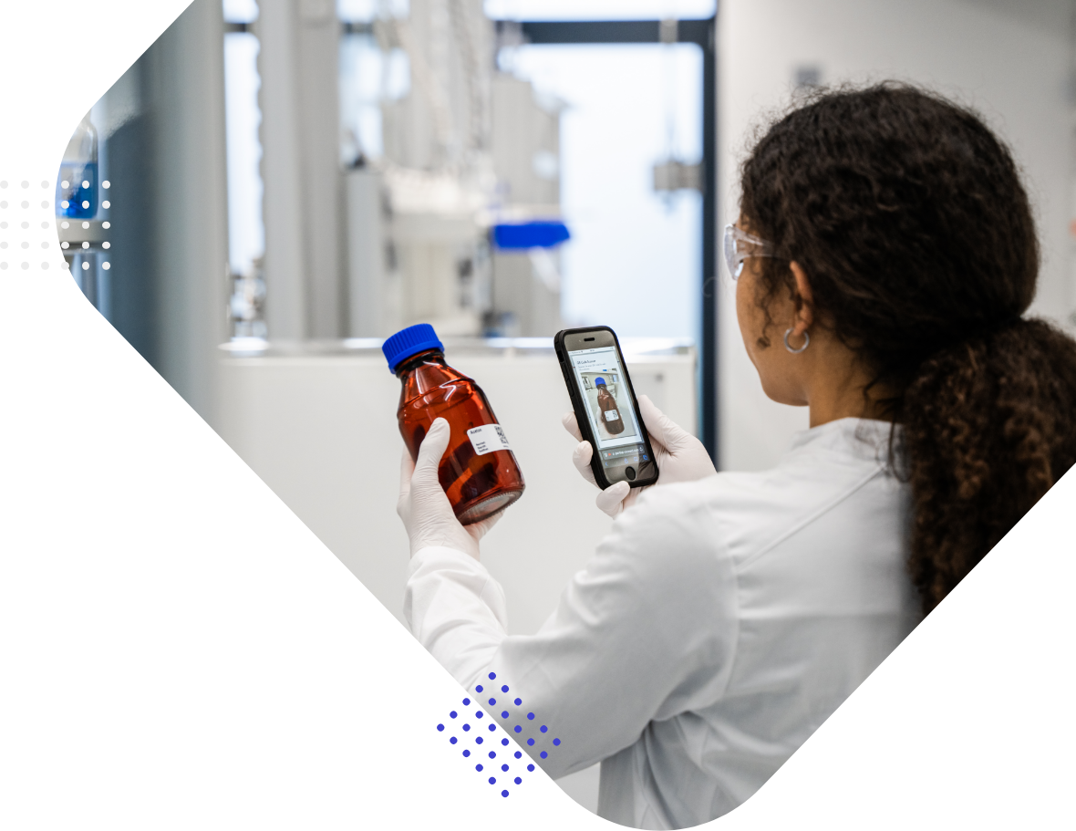 Eine Labormitarbeiterin hält ein rotes Fläschchen und schaut auf ihr Smartphone.