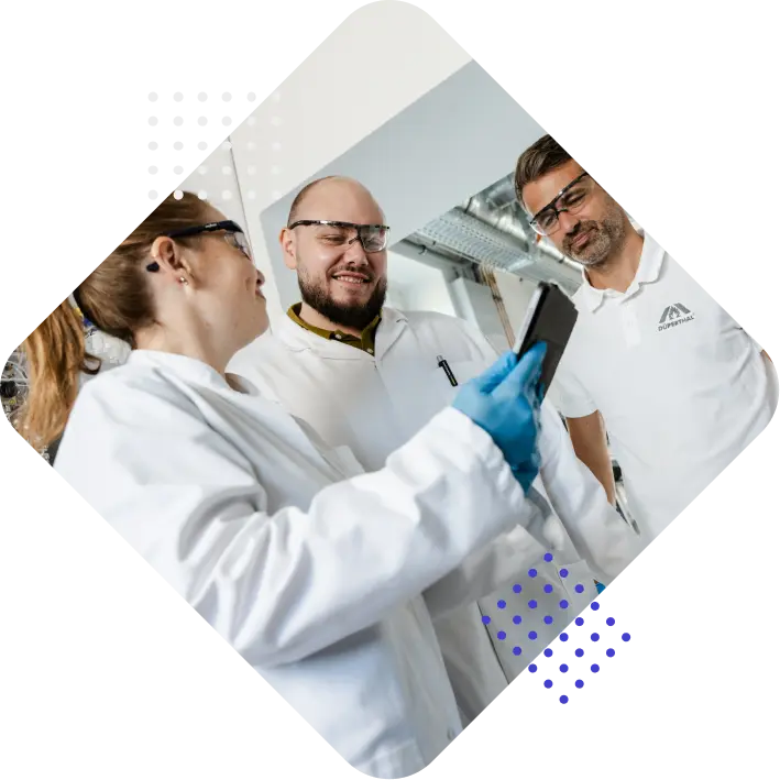 Three laboratory professionals in white coats and safety glasses are gathered around a smartphone, smiling and engaging in conversation.