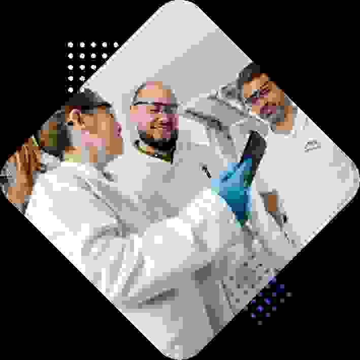 Three laboratory professionals in white coats and safety glasses are gathered around a smartphone, smiling and engaging in conversation.