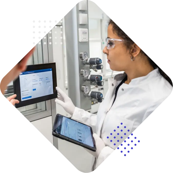 A woman in a lab coat examines data on a digital tablet and touchscreen display in a laboratory setting, with equipment and gauges visible in the background.