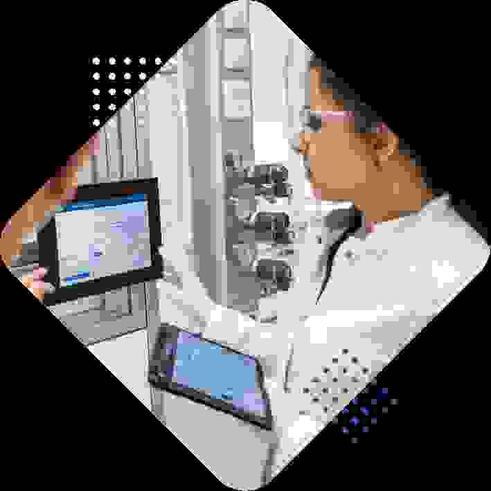A woman in a lab coat examines data on a digital tablet and touchscreen display in a laboratory setting, with equipment and gauges visible in the background.