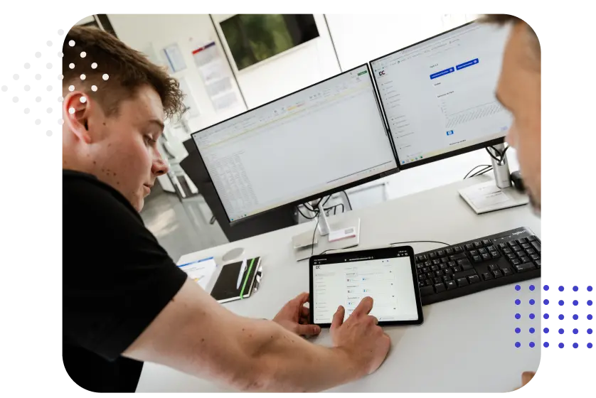 Two individuals are discussing data displayed on a tablet while sitting at a desk with two computer monitors. One person is pointing at the tablet screen, which shows an interface, and there are office supplies on the desk.