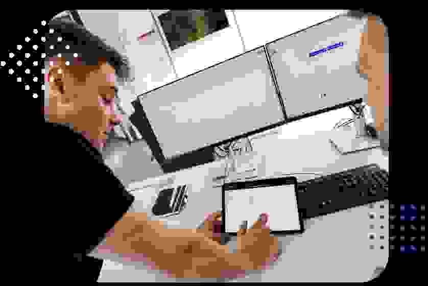 Two individuals are discussing data displayed on a tablet while sitting at a desk with two computer monitors. One person is pointing at the tablet screen, which shows an interface, and there are office supplies on the desk.