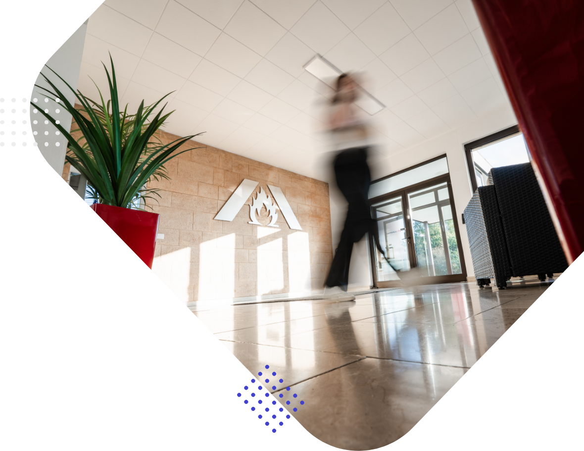 A blurred figure walks in a lobby with a large plant and a wall sign featuring a flame logo. The space has large windows and modern decor.