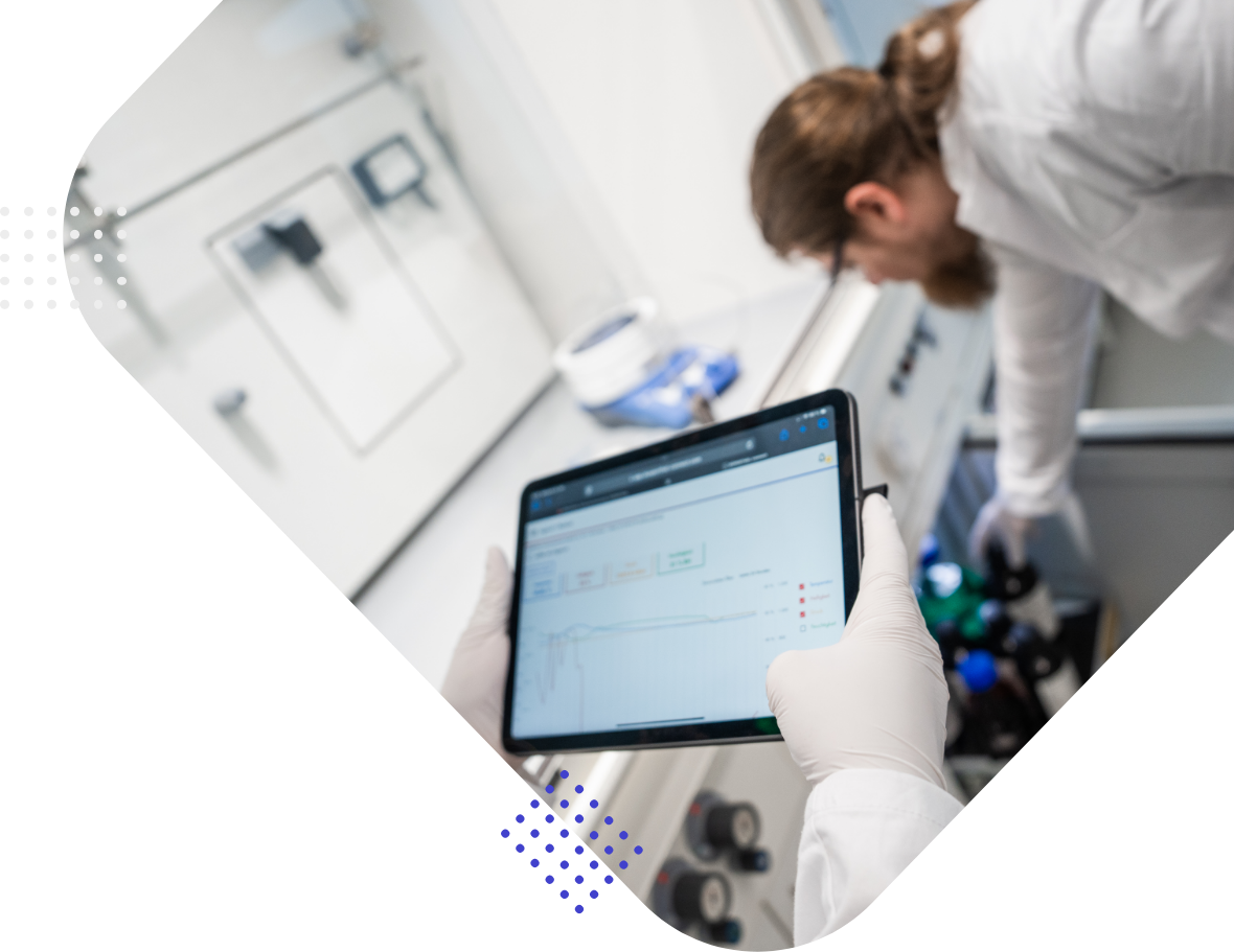 A laboratory setting showing a person in a white lab coat and gloves using a tablet to display data, while another individual is working with equipment in the background.