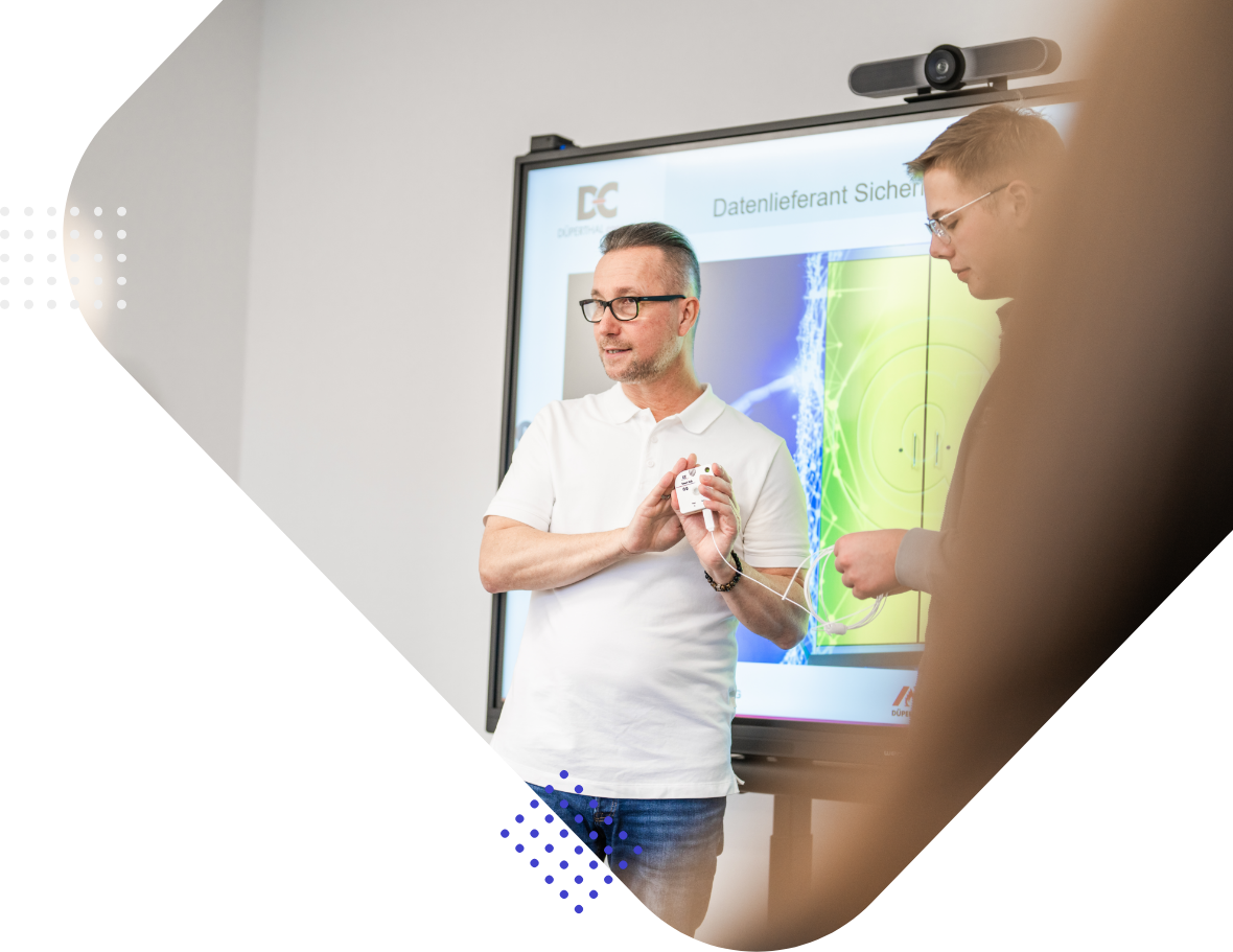 Two men are engaged in a discussion during a presentation. One man, wearing a white shirt, holds a device and gestures, while the other man, wearing glasses, is interacting with him. A digital screen behind them displays graphical information.