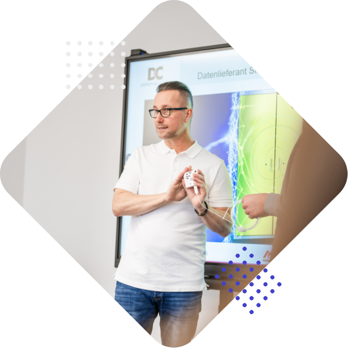 A man in a white polo shirt is presenting in front of a screen, holding a small device. The screen displays graphs and text in German.
