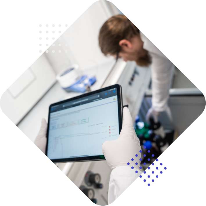 Eine Person mit weißen Handschuhen hält ein Tablet, auf dem Daten angezeigt werden, während im Hintergrund eine andere Person in einem Labor arbeitet.