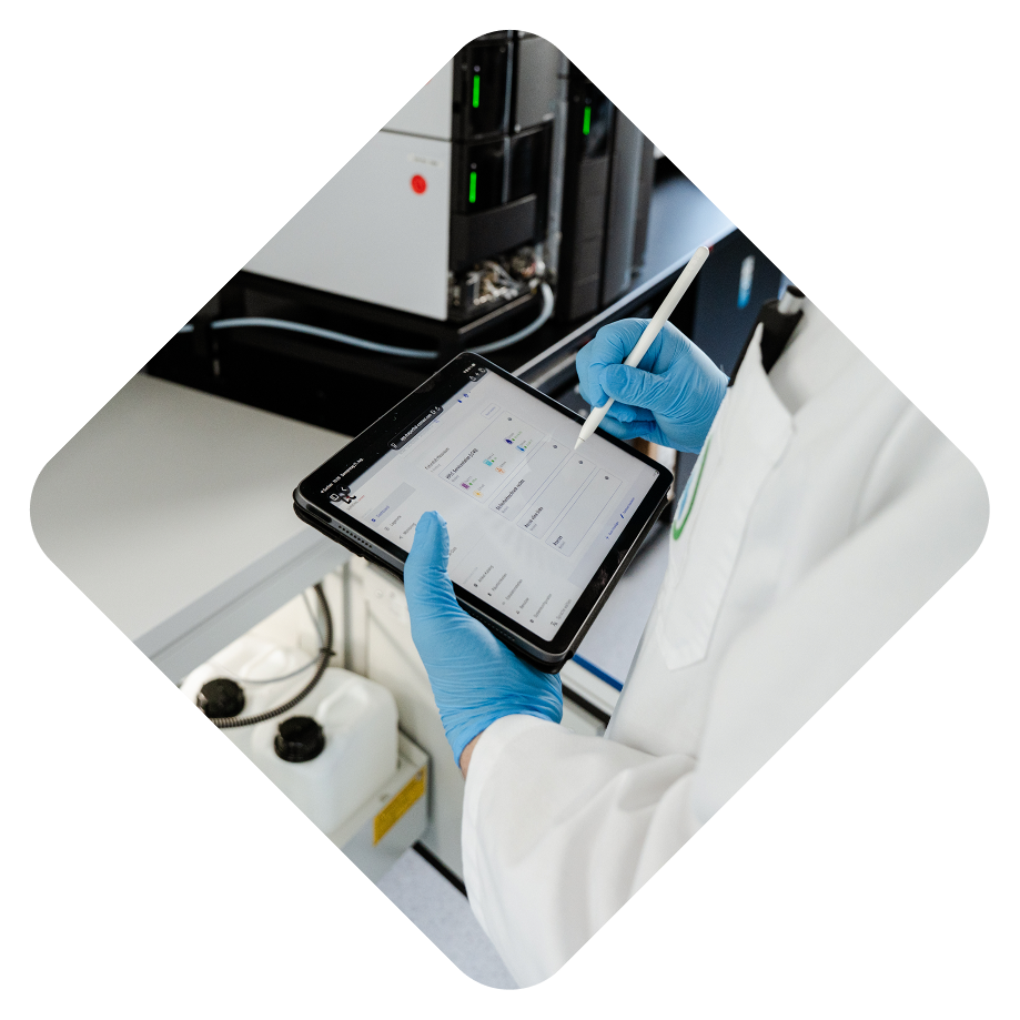 Ein Laborant in weißem Kittel und blauen Handschuhen benutzt ein Tablet mit einem Stylus, während er an einem Tisch in einem Labor arbeitet.