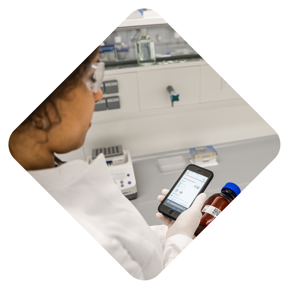 Eine Person in einem Labor hält ein Smartphone und eine Flasche. Sie trägt Handschuhe und schaut auf das Handy.